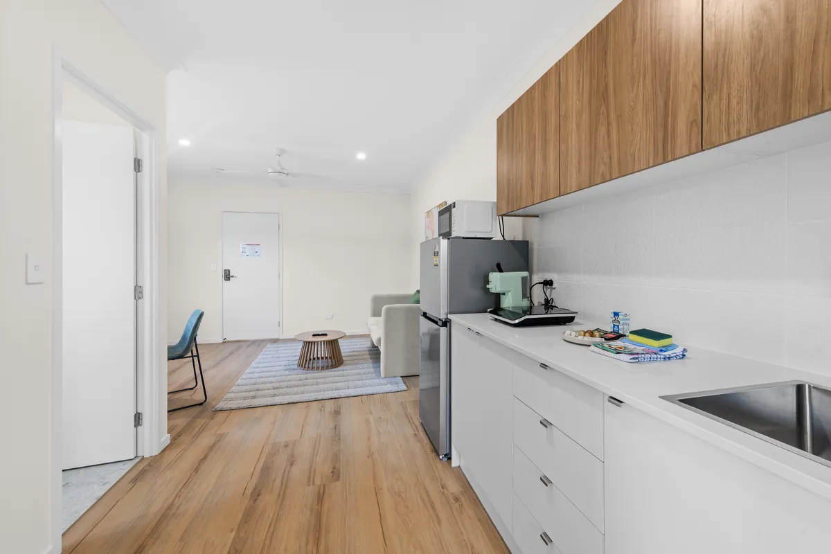 Kitchenette in a Brisbane rooming house with white cabinets, timber overhead cupboards and a fridge